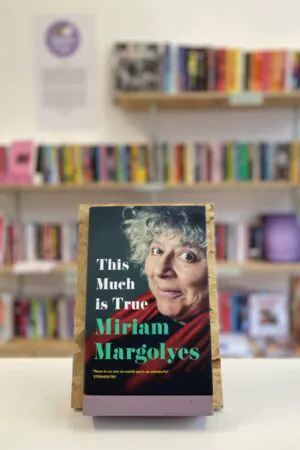 Cymraeg: Copi o 'This Much Is True' yn sefyll ar stondin llyfrau, tu blaen silffoedd o lyfrau yn y cefndir. | English: A copy of 'This Much Is True' sits on a stand in front of multiple shelves of other books.