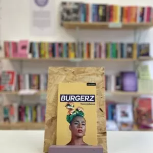 Cymraeg: Copi o 'Burgerz' yn sefyll ar stondin llyfrau, tu blaen silffoedd o lyfrau yn y cefndir. English: A copy of 'Burgerz' sitson a stand in front of multiple shelves of other books.
