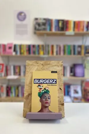 Cymraeg: Copi o 'Burgerz' yn sefyll ar stondin llyfrau, tu blaen silffoedd o lyfrau yn y cefndir. English: A copy of 'Burgerz' sitson a stand in front of multiple shelves of other books.