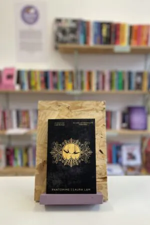 Cymraeg: Copi o 'Pantomime' yn sefyll ar stondin llyfrau, tu blaen silffoedd o lyfrau yn y cefndir. English: A copy of 'Pantomime' sitson a stand in front of multiple shelves of other books.
