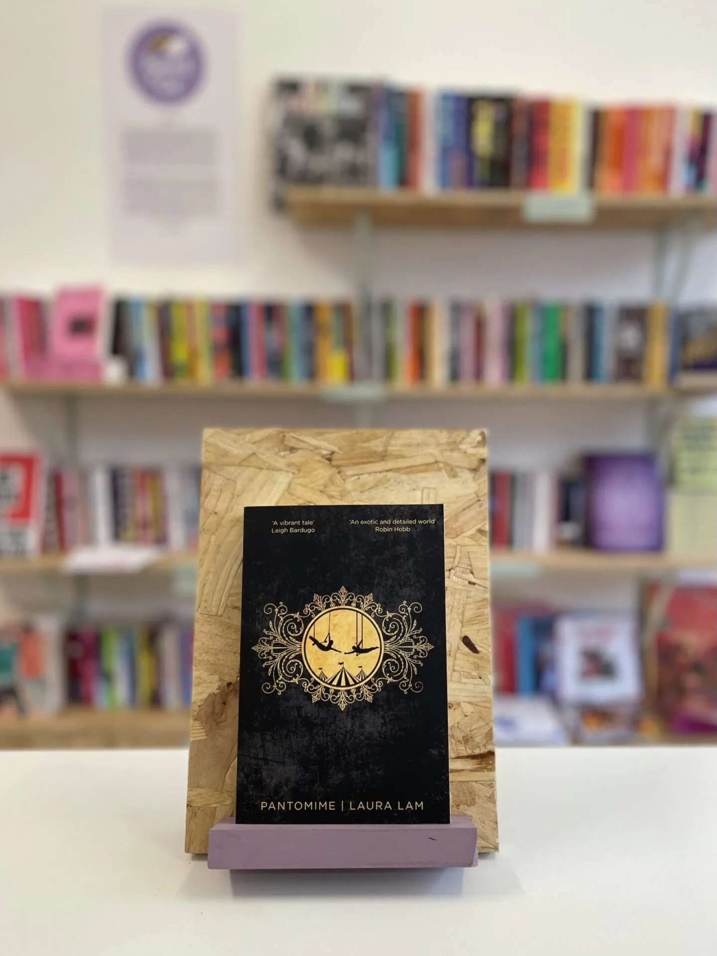Cymraeg: Copi o 'Pantomime' yn sefyll ar stondin llyfrau, tu blaen silffoedd o lyfrau yn y cefndir. English: A copy of 'Pantomime' sitson a stand in front of multiple shelves of other books.