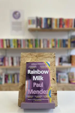 Cymraeg: Copi o 'Rainbow Milk' yn sefyll ar stondin llyfrau, tu blaen silffoedd o lyfrau yn y cefndir. | English: A copy of 'Rainbow Milk' sits on a stand in front of multiple shelves of other books.
