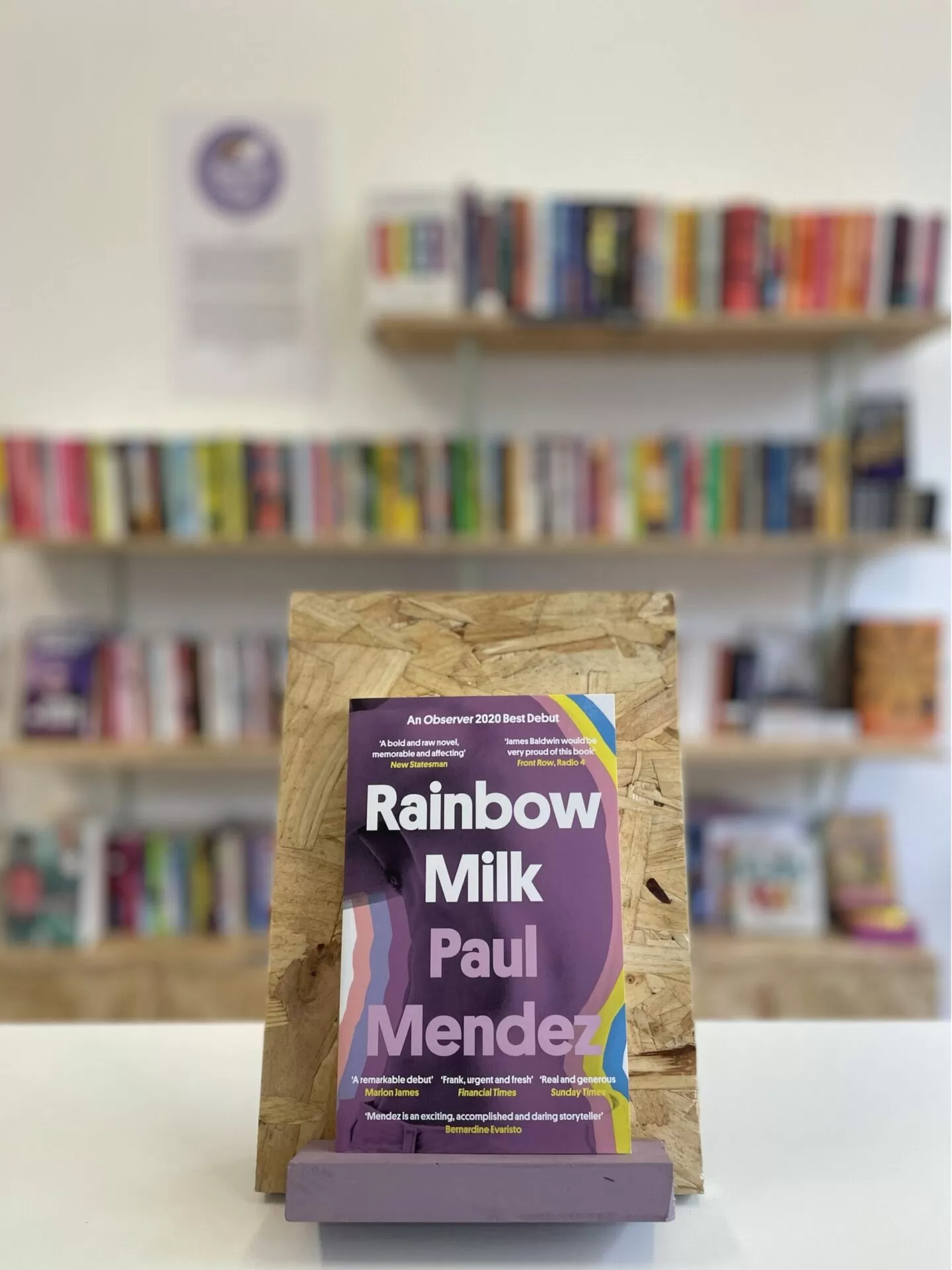 Cymraeg: Copi o 'Rainbow Milk' yn sefyll ar stondin llyfrau, tu blaen silffoedd o lyfrau yn y cefndir. | English: A copy of 'Rainbow Milk' sits on a stand in front of multiple shelves of other books.