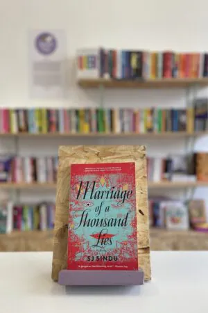 A copy of 'Marriage of a Thousand Lies' sits on a stand in front of multiple shelves of other books.
