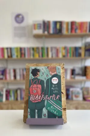 A copy of 'Heartstopper vol. 1' sits on a stand in front of multiple shelves of other books.