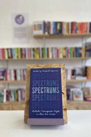 Cymraeg: Copi o 'Spectrums' yn sefyll ar stondin llyfrau, tu blaen silffoedd o lyfrau yn y cefndir. | English: A copy of 'Spectrums' sits on a stand in front of multiple shelves of other books.