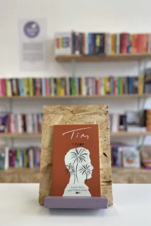 Cymraeg: Copi o 'Y Pump: Tim' yn sefyll ar stondin llyfrau, tu blaen silffoedd o lyfrau yn y cefndir. | English: A copy of 'Y Pump: Tim' sits on a stand in front of multiple shelves of other books.
