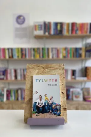 Cymraeg: Copi o 'Tylwyth' yn sefyll ar stondin llyfrau, tu blaen silffoedd o lyfrau yn y cefndir. | English: A copy of 'Tylwyth' sits on a stand in front of multiple shelves of other books.