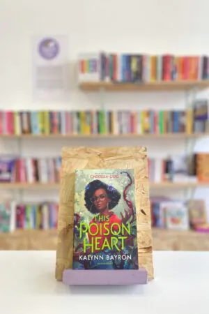 Cymraeg: Copi o 'The Poison Heart' yn sefyll ar stondin llyfrau, tu blaen silffoedd o lyfrau yn y cefndir. | English: A copy of 'The Poison Heart' sits on a stand in front of multiple shelves of other books.