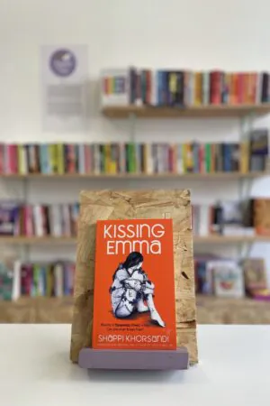 Cymraeg: Copi o 'Kissing Emma' yn sefyll ar stondin llyfrau, tu blaen silffoedd o lyfrau yn y cefndir. | English: A copy of 'Kissing Emma' sits on a stand in front of multiple shelves of other books.