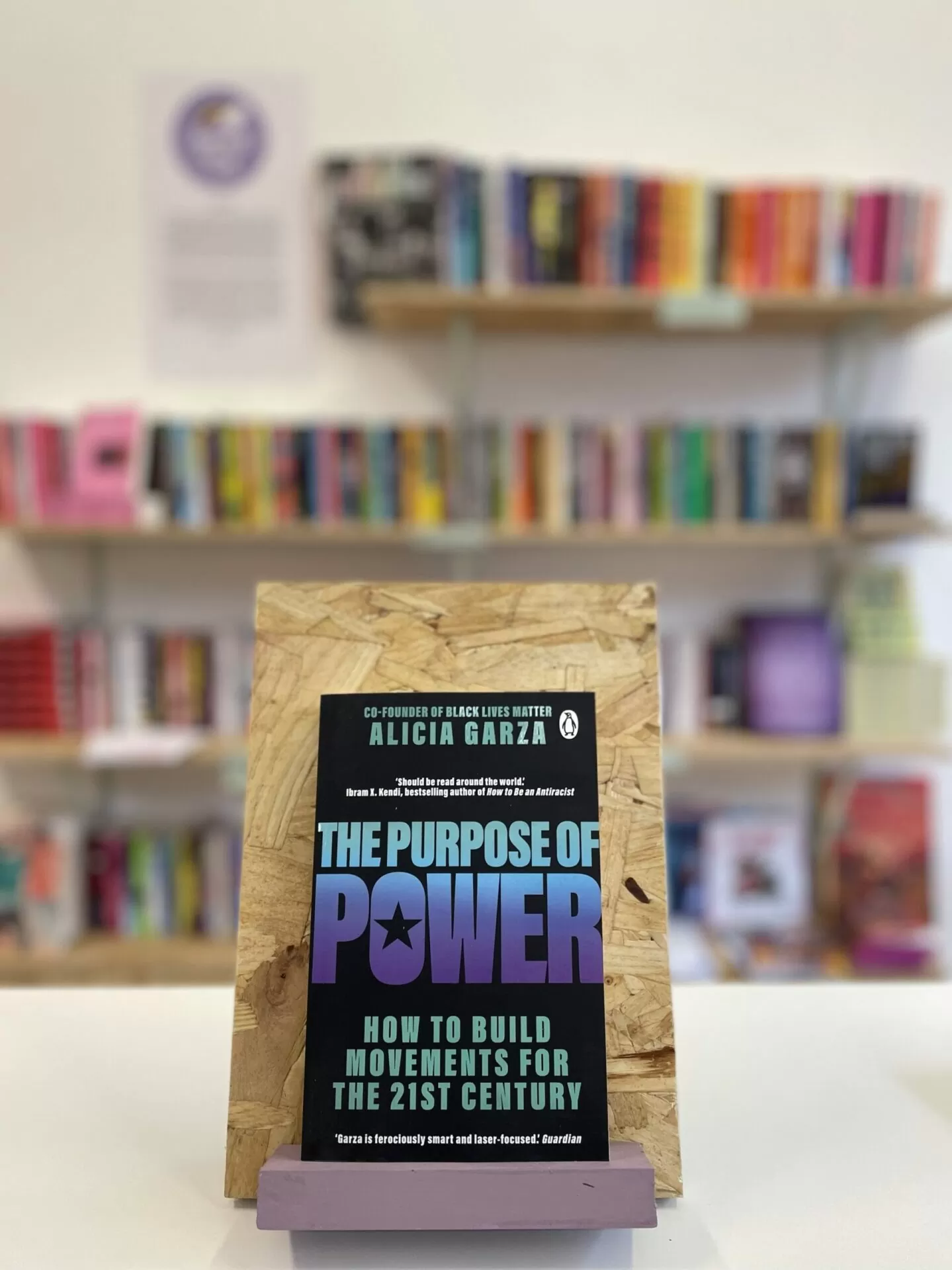 Cymraeg: Copi o 'The Purpose of Power' yn sefyll ar stondin llyfrau, tu blaen silffoedd o lyfrau yn y cefndir. | English: A copy of 'The Purpose of Power' sits on a stand in front of multiple shelves of other books.