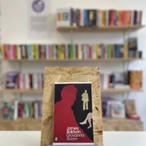 Cymraeg: Copi o 'Giovanni's Room' yn sefyll ar stondin llyfrau, tu blaen silffoedd o lyfrau yn y cefndir. | English: A copy of 'Giovanni's Room' sits on a stand in front of multiple shelves of other books.