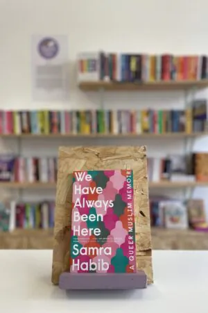 Cymraeg: Copi o 'We Have Always Been Here' yn sefyll ar stondin llyfrau, tu blaen silffoedd o lyfrau yn y cefndir. | English: A copy of 'We Have Always Been Here' sits on a stand in front of multiple shelves of other books.