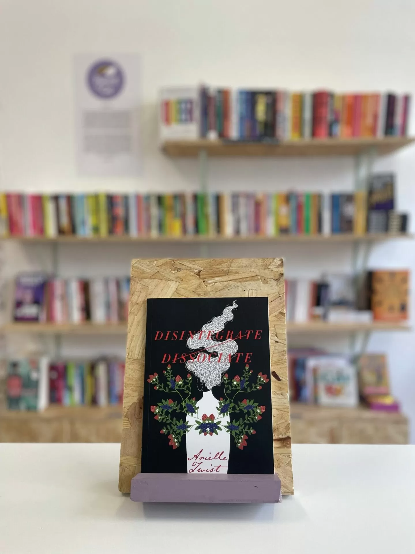 A copy of 'Disintegrate/Dissociate' sits on a stand in front of multiple shelves of other books.