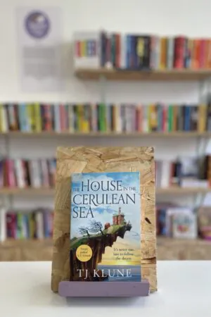 Cymraeg: Copi o 'The House in the Cerulean Sea' yn sefyll ar stondin llyfrau, tu blaen silffoedd o lyfrau yn y cefndir. | English: A copy of 'The House in the Cerulean Sea' sits on a stand in front of multiple shelves of other books.