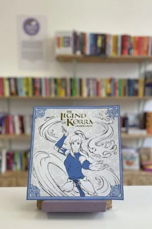 Cymraeg: Copi o 'The Legend of Korra Coloring Book' yn sefyll ar stondin llyfrau, tu blaen silffoedd o lyfrau yn y cefndir. English: A copy of 'The Legend of Korra Coloring Book' sits on a stand in front of multiple shelves of other books.