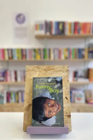 Cymraeg: Copi o 'Funny Boy' yn sefyll ar stondin llyfrau, tu blaen silffoedd o lyfrau yn y cefndir. | English: A copy of 'Funny Boy' sits on a stand in front of multiple shelves of other books.