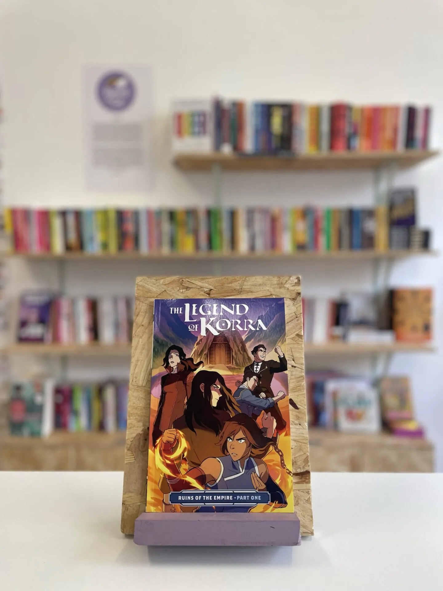 Cymraeg: Copi o 'The Legend of Korra: Ruins of the Empire, Part One' yn sefyll ar stondin llyfrau, tu blaen silffoedd o lyfrau yn y cefndir. | English: A copy of 'The Legend of Korra: Ruins of the Empire, Part One' sits on a stand in front of multiple shelves of other books.