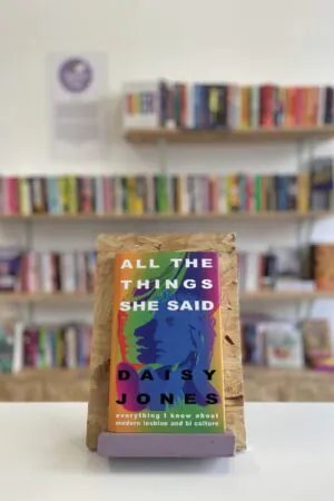Cymraeg: Copi o 'All the Things She Said' yn sefyll ar stondin llyfrau, tu blaen silffoedd o lyfrau yn y cefndir. | English: A copy of 'All the Things She Said' sits on a stand in front of multiple shelves of other books.