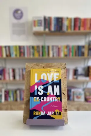 Cymraeg: Copi o 'Love Is An Ex-Country' yn sefyll ar stondin llyfrau, tu blaen silffoedd o lyfrau yn y cefndir. | English: A copy of 'Love Is An Ex-Country' sits on a stand in front of multiple shelves of other books.