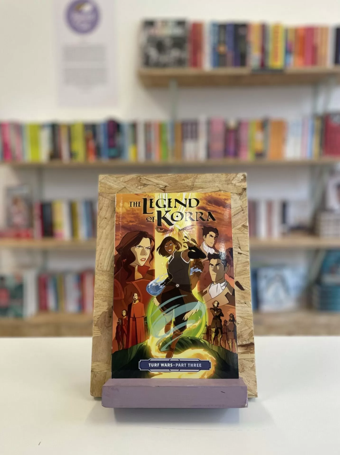 Cymraeg: Copi o 'The Legend of Korra: Turf Wars, Part Three' yn sefyll ar stondin llyfrau, tu blaen silffoedd o lyfrau yn y cefndir. | English: A copy of 'The Legend of Korra: Turf Wars, Part Three' sits on a stand in front of multiple shelves of other books.