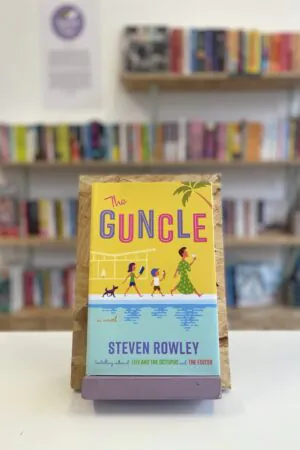Cymraeg: Copi o 'The Guncle' yn sefyll ar stondin llyfrau, tu blaen silffoedd o lyfrau yn y cefndir. | English: A copy of 'The Guncle' sits on a stand in front of multiple shelves of other books.