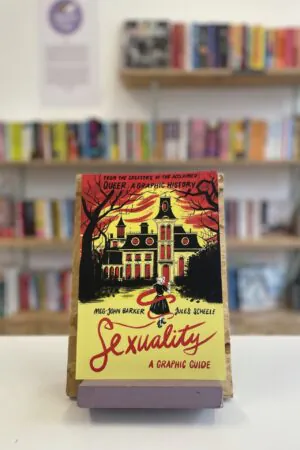 Cymraeg: Copi o 'Sexuality: A Graphic Guide' yn sefyll ar stondin llyfrau, tu blaen silffoedd o lyfrau yn y cefndir. | English: A copy of 'Sexuality: A Graphic Guide' sits on a stand in front of multiple shelves of other books.