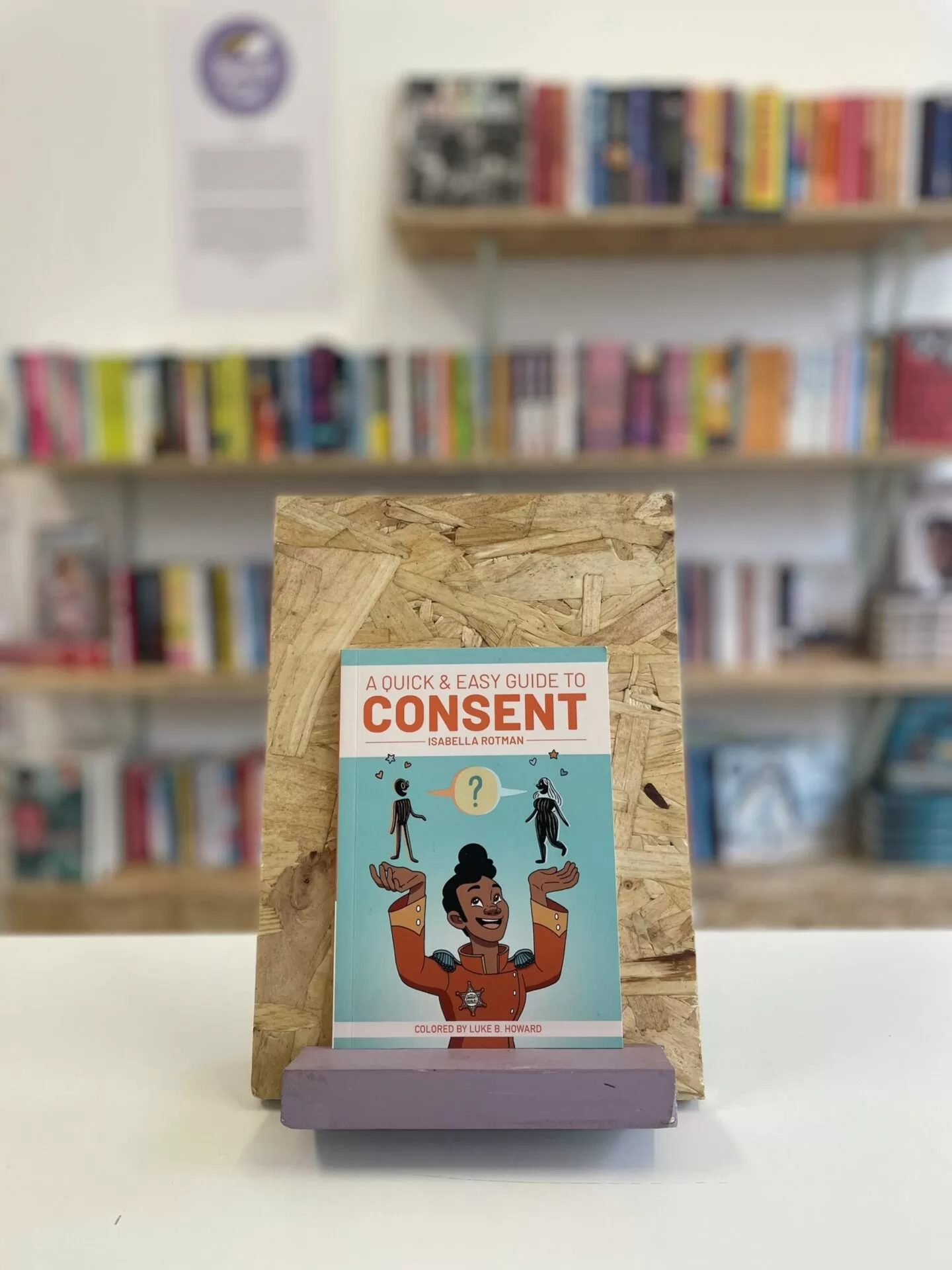 Cymraeg: Copi o 'A Quick & Easy Guide to Consent' yn sefyll ar stondin llyfrau, tu blaen silffoedd o lyfrau yn y cefndir. | English: A copy of 'A Quick & Easy Guide to Consent' sits on a stand in front of multiple shelves of other books.