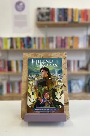 Cymraeg: Copi o 'The Legend of Korra: Ruins of the Empire, Part Two' yn sefyll ar stondin llyfrau, tu blaen silffoedd o lyfrau yn y cefndir. | English: A copy of 'The Legend of Korra: Ruins of the Empire, Part Two' sits on a stand in front of multiple shelves of other books.