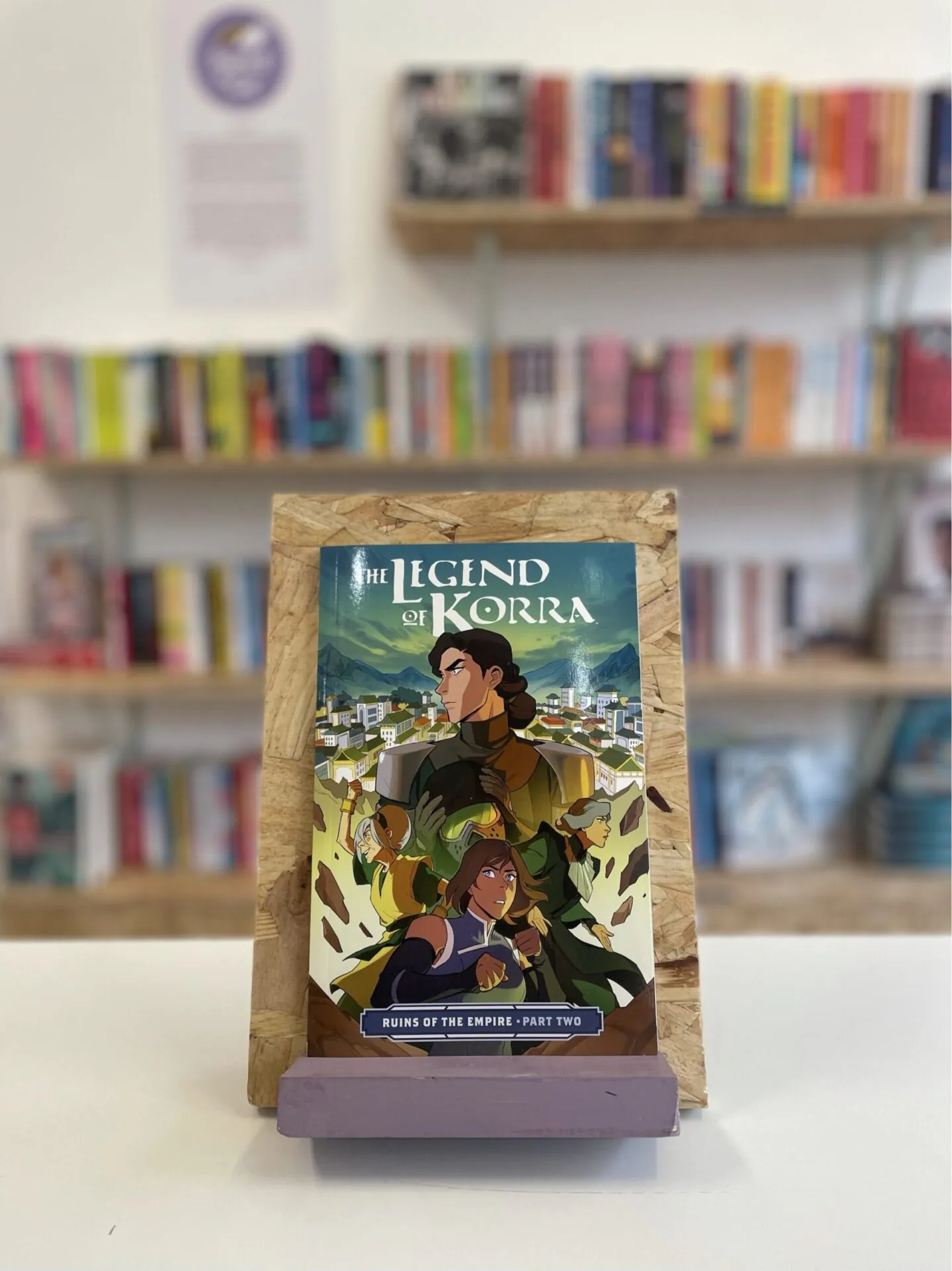 Cymraeg: Copi o 'The Legend of Korra: Ruins of the Empire, Part Two' yn sefyll ar stondin llyfrau, tu blaen silffoedd o lyfrau yn y cefndir. | English: A copy of 'The Legend of Korra: Ruins of the Empire, Part Two' sits on a stand in front of multiple shelves of other books.