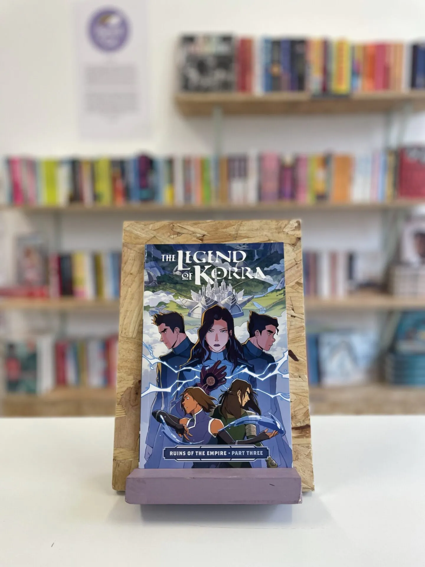 Cymraeg: Copi o 'The Legend of Korra: Ruins of the Empire, Part Three' yn sefyll ar stondin llyfrau, tu blaen silffoedd o lyfrau yn y cefndir. | English: A copy of 'The Legend of Korra: Ruins of the Empire, Part Three' sits on a stand in front of multiple shelves of other books.