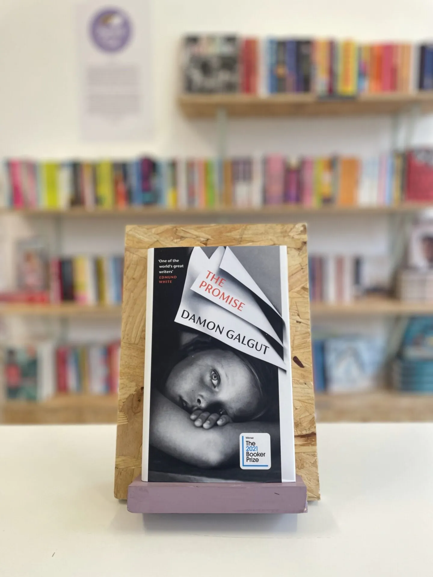 Cymraeg: Copi o 'The Promise yn sefyll ar stondin llyfrau, tu blaen silffoedd o lyfrau yn y cefndir. | English: A copy of 'The Promise' sits on a stand in front of multiple shelves of other books.