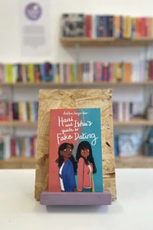 Cymraeg: Copi o 'Hani and Ishu's Guide to Fake Dating' yn sefyll ar stondin llyfrau, tu blaen silffoedd o lyfrau yn y cefndir. | English: A copy of 'Hani and Ishu's Guide to Fake Dating' sits on a stand in front of multiple shelves of other books.