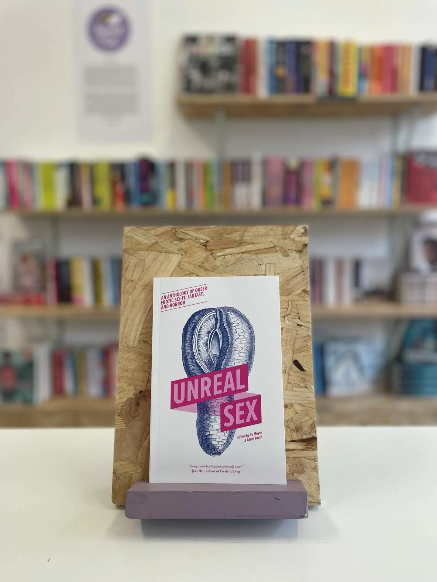 Cymraeg: Copi o 'Unreal Sex' yn sefyll ar stondin llyfrau, tu blaen silffoedd o lyfrau yn y cefndir. | English: A copy of 'Unreal Sex' sits on a stand in front of multiple shelves of other books.