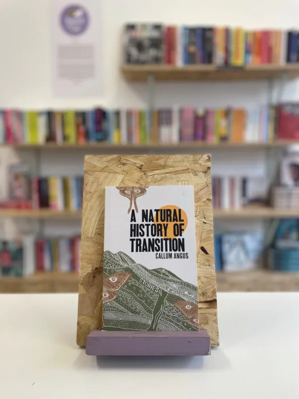 A copy of 'A Natural History of Transition' sits on a stand in front of multiple shelves of other books.