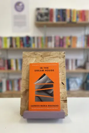 Cymraeg: Copi o 'In the Dream House' yn sefyll ar stondin llyfrau, tu blaen silffoedd o lyfrau yn y cefndir. | English: A copy of 'In the Dream House' sits on a stand in front of multiple shelves of other books.