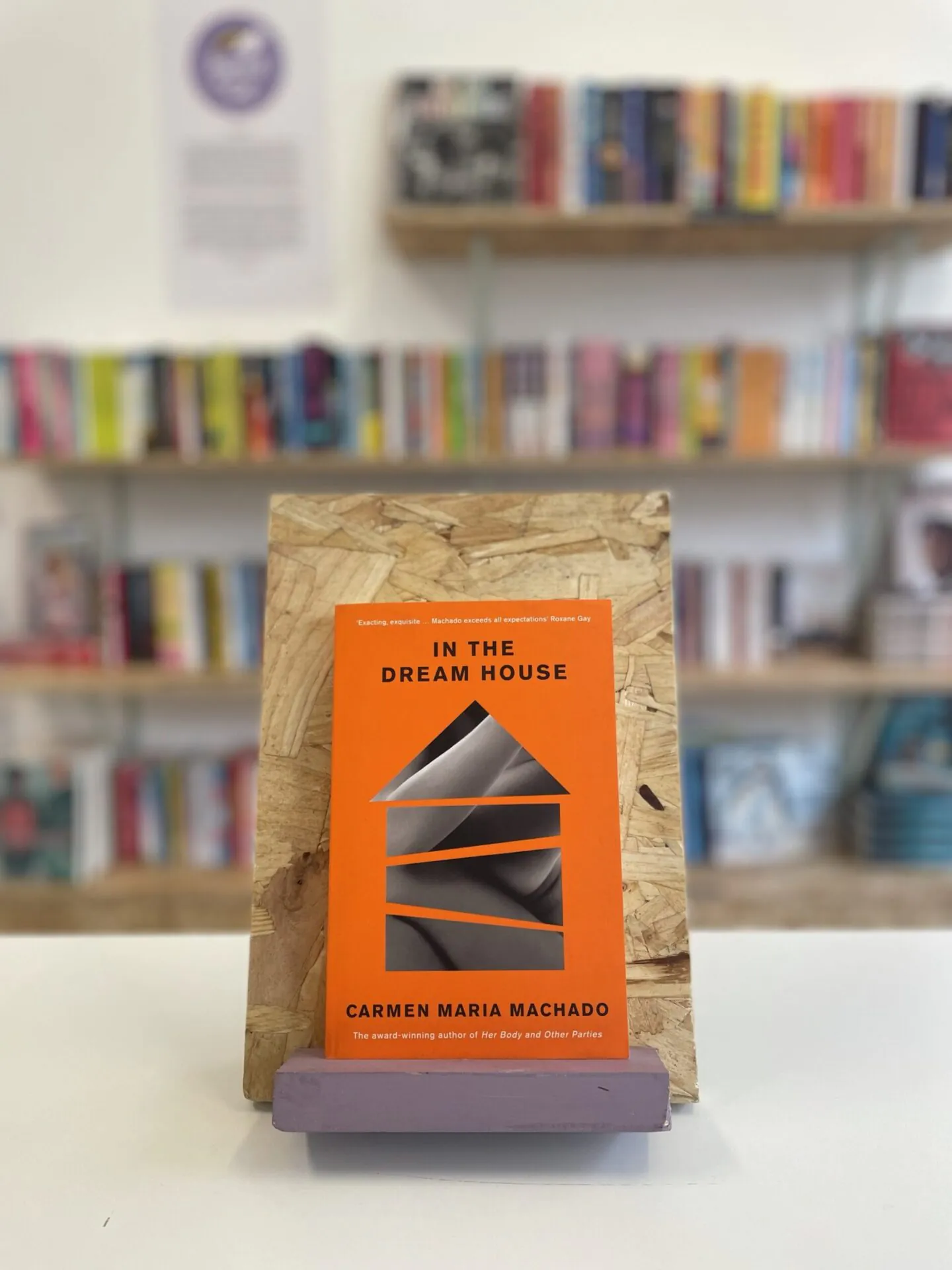 Cymraeg: Copi o 'In the Dream House' yn sefyll ar stondin llyfrau, tu blaen silffoedd o lyfrau yn y cefndir. | English: A copy of 'In the Dream House' sits on a stand in front of multiple shelves of other books.