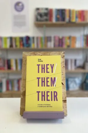 Cymraeg: Copi o 'They/Them/Their' yn sefyll ar stondin llyfrau, tu blaen silffoedd o lyfrau yn y cefndir. | English: A copy of 'They/Them/Their' sits on a stand in front of multiple shelves of other books.
