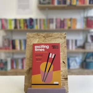 Cymraeg: Copi o 'Exciting Times' yn sefyll ar stondin llyfrau, tu blaen silffoedd o lyfrau yn y cefndir. | English: A copy of 'Exciting Times' sits on a stand in front of multiple shelves of other books.