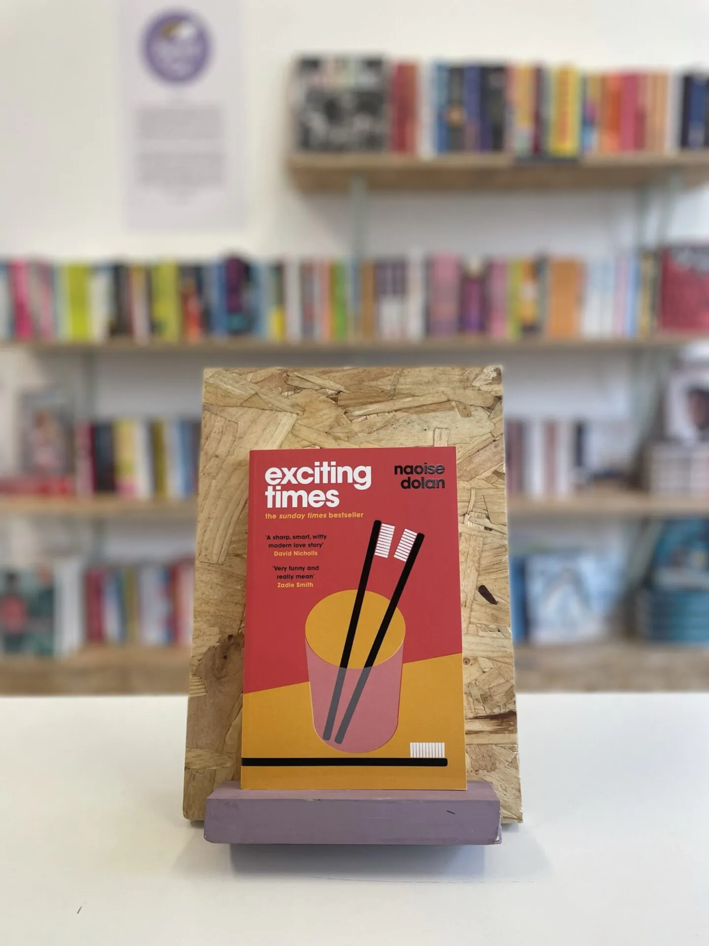 Cymraeg: Copi o 'Exciting Times' yn sefyll ar stondin llyfrau, tu blaen silffoedd o lyfrau yn y cefndir. | English: A copy of 'Exciting Times' sits on a stand in front of multiple shelves of other books.