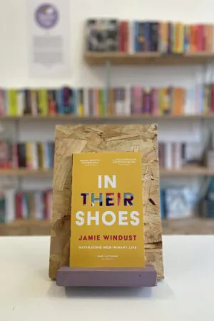 Cymraeg: Copi o 'In Their Shoes' yn sefyll ar stondin llyfrau, tu blaen silffoedd o lyfrau yn y cefndir. | English: A copy of 'In Their Shoes' sits on a stand in front of multiple shelves of other books.