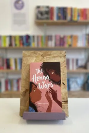Cymraeg: Copi o 'The Henna Wars' yn sefyll ar stondin llyfrau, tu blaen silffoedd o lyfrau yn y cefndir. | English: A copy of 'The Henna Wars' sits on a stand in front of multiple shelves of other books.