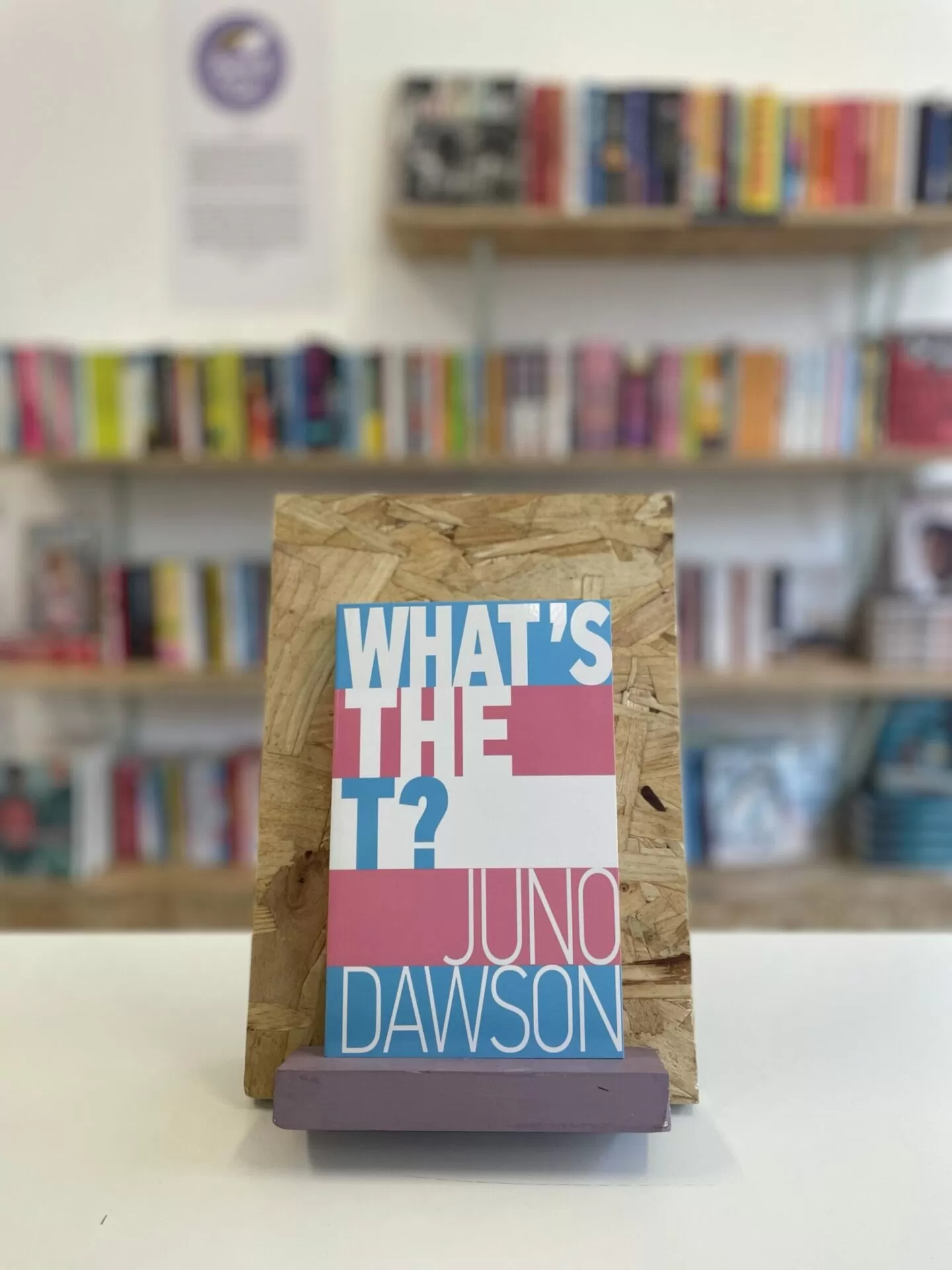 A copy of 'What's the T?' sits on a stand in front of multiple shelves of other books.