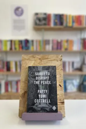 Cymraeg: Copi o 'Drag Queen of Scots' yn sefyll ar stondin llyfrau, tu blaen silffoedd o lyfrau yn y cefndir. | English: A copy of 'Drag Queen of Scots' sits on a stand in front of multiple shelves of other books.