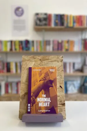 Cymraeg: Copi o 'The Normal Heart' yn sefyll ar stondin llyfrau, tu blaen silffoedd o lyfrau yn y cefndir. | English: A copy of 'The Normal Heart' sits on a stand in front of multiple shelves of other books.