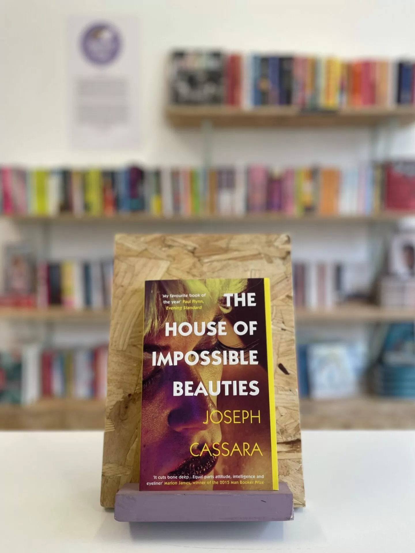 Cymraeg: Copi o 'The House of Impossible Beauties' yn sefyll ar stondin llyfrau, tu blaen silffoedd o lyfrau yn y cefndir. | English: A copy of 'The House of Impossible Beauties' sits on a stand in front of multiple shelves of other books.