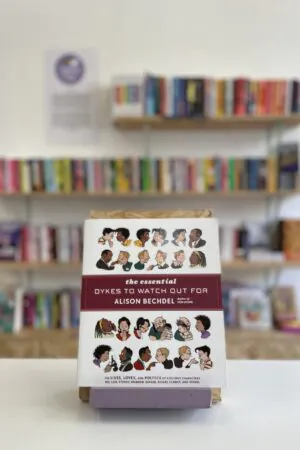 Cymraeg: Copi o 'The Essential Dykes to Watch Our For' yn sefyll ar stondin llyfrau, tu blaen silffoedd o lyfrau yn y cefndir. | English: A copy of 'The Essential Dykes to Watch Our For' sits on a stand in front of multiple shelves of other books.