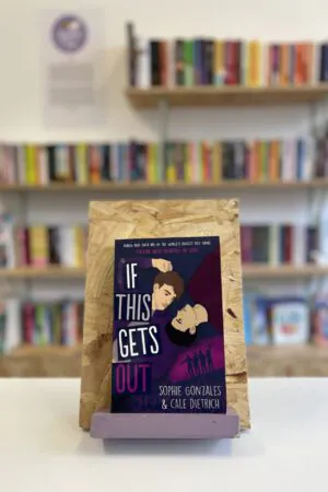 Cymraeg: Copi o 'If This Gets Out' yn sefyll ar stondin llyfrau, tu blaen silffoedd o lyfrau yn y cefndir. | English: A copy of 'If This Gets Out' sits on a stand in front of multiple shelves of other books.