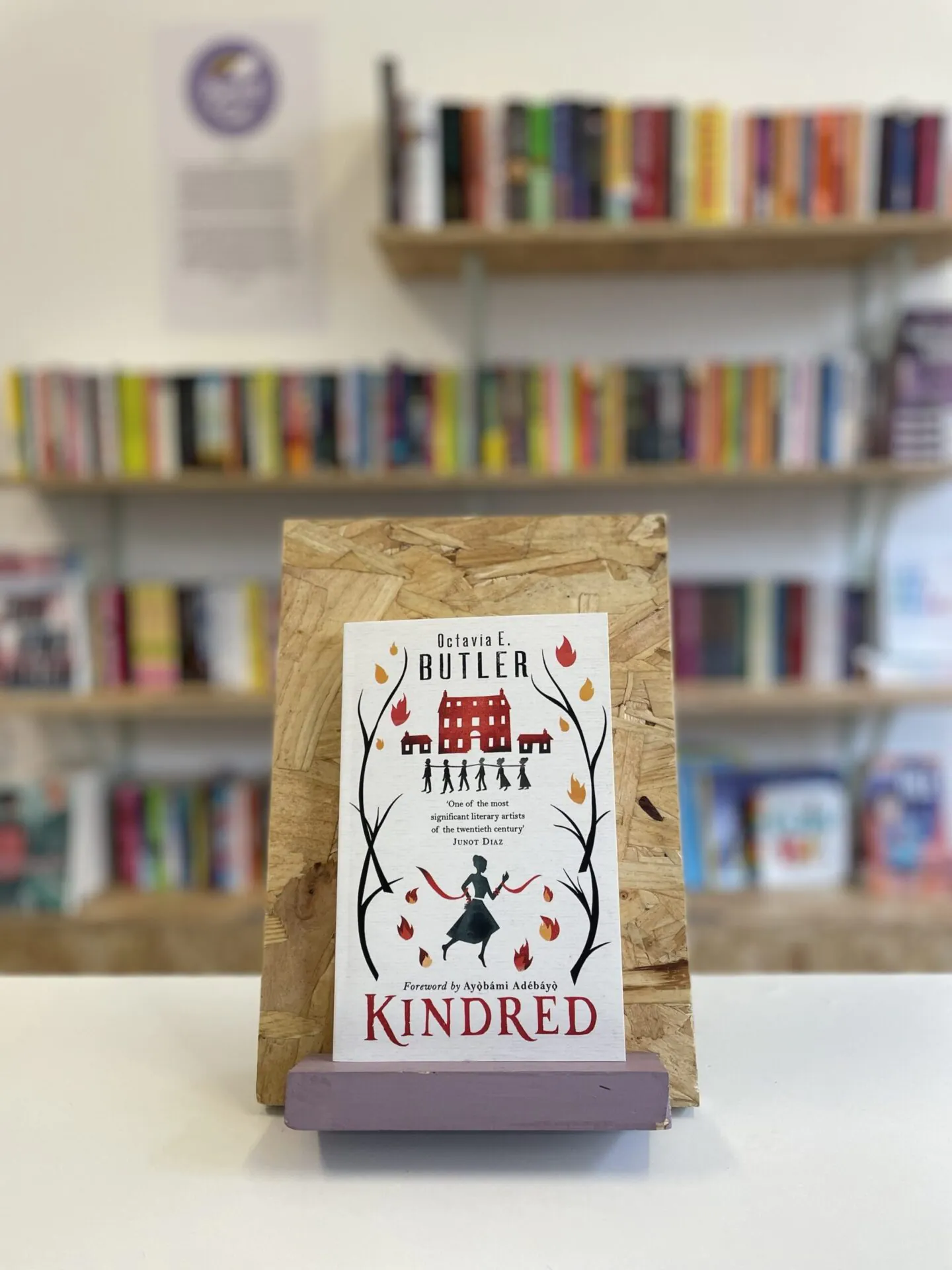 Cymraeg: Copi o 'Kindred' yn sefyll ar stondin llyfrau, tu blaen silffoedd o lyfrau yn y cefndir. | English: A copy of 'Kindred' sits on a stand in front of multiple shelves of other books.