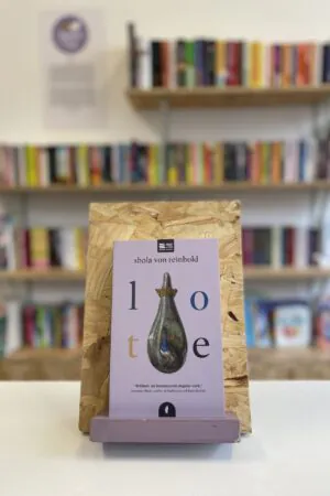 Cymraeg: Copi o 'Lote' yn sefyll ar stondin llyfrau, tu blaen silffoedd o lyfrau yn y cefndir. | English: A copy of 'Lote' sits on a stand in front of multiple shelves of other books.